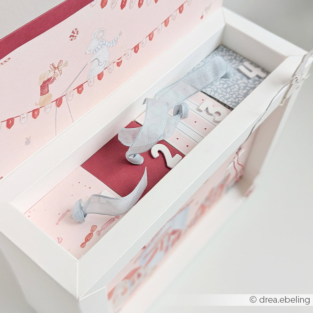 Eine offene, weiße Geschenkbox, in der ein nummerierter Adventskalender mit festlichen Designpapieren zu sehen ist.