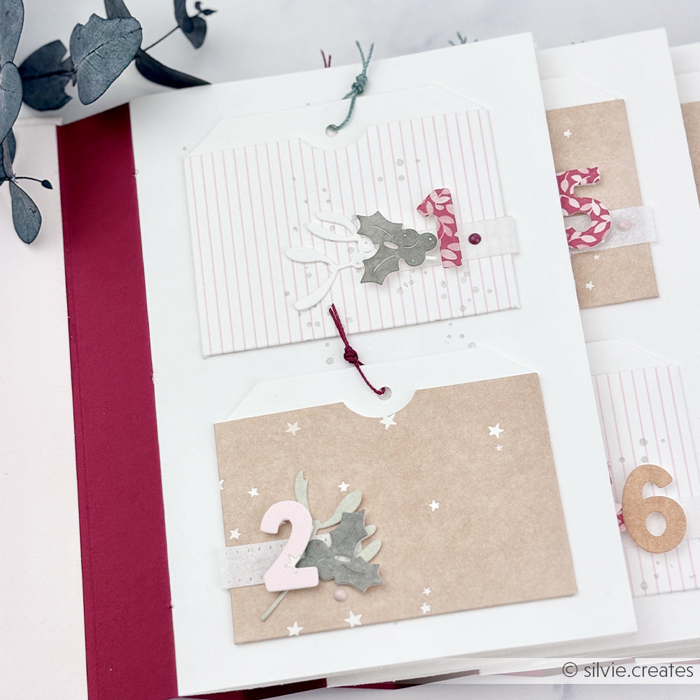 Handgefertigte Adventskalenderseiten mit nummerierten Anhängern und festlichen Verzierungen aus Designpapier.