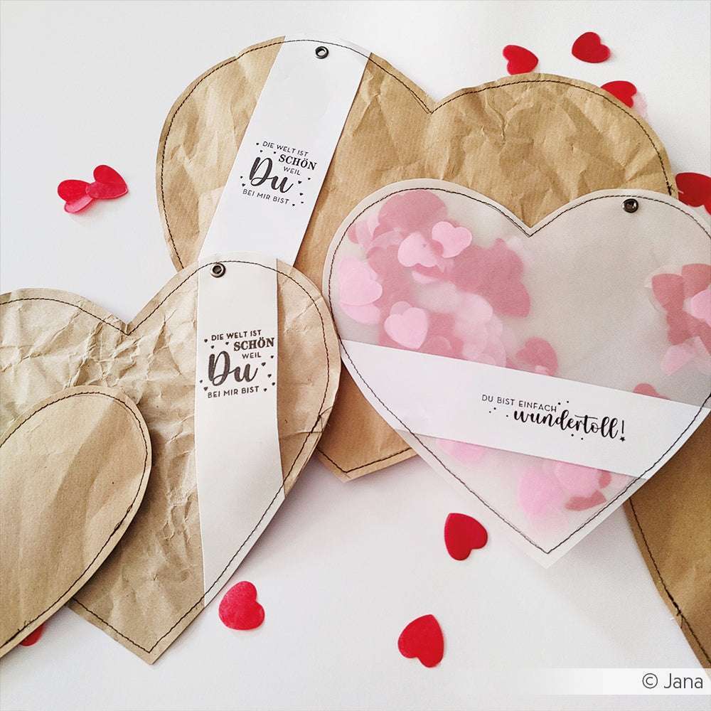 Drei herzförmige Papierbasteleien von Alexandra Renke, einige mit durchsichtigen Fronten und rosafarbenen Herzen, sind von rotem Konfetti umgeben - ideal für Papierprojekte oder in Kombination mit Ihrem Lieblingsholzstempel.