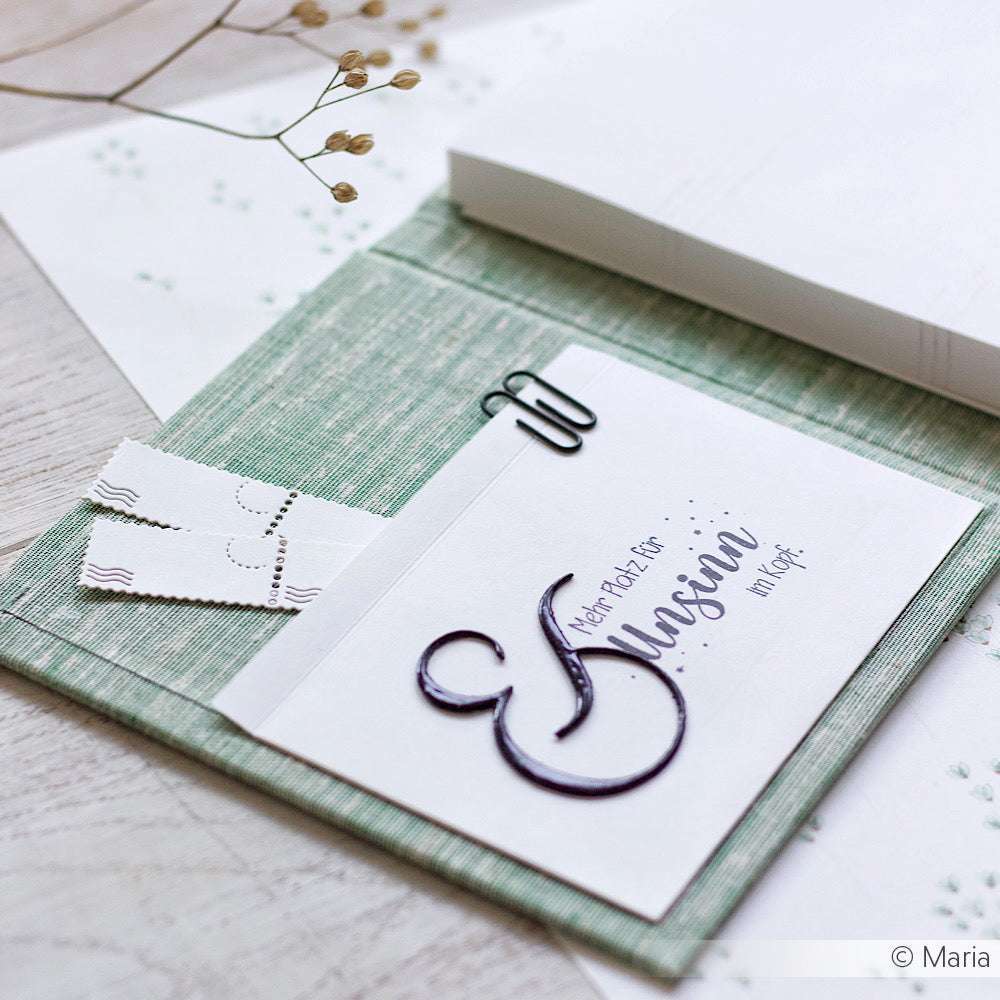 Eine handgeschriebene Einladung mit Holzstempel-Akzenten und einer Büroklammer ruht auf einer strukturierten grünen Mappe von Alexandra Renke, perfekt für Papierprojekte.