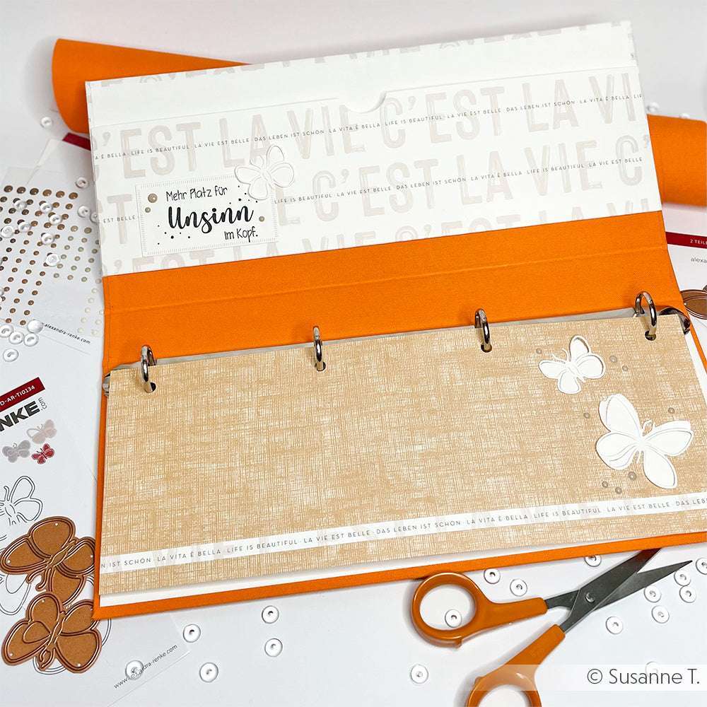 Geöffnete orangefarbene Mappe mit Scrapbook-Materialien, Schere, Alexandra Renke Holzstempel, Stickern und Papierschmetterlingen auf einer weißen Fläche - perfekt für kreative Papierprojekte.