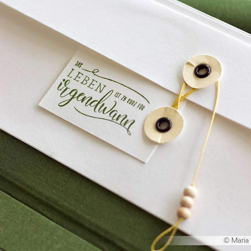Alexandra Renke weißer Umschlag mit Kordelverschluss auf grünem Stoff, ideal für Papierprojekte. Tag sagt "Das Leben ist zu kurz für irgendwann.