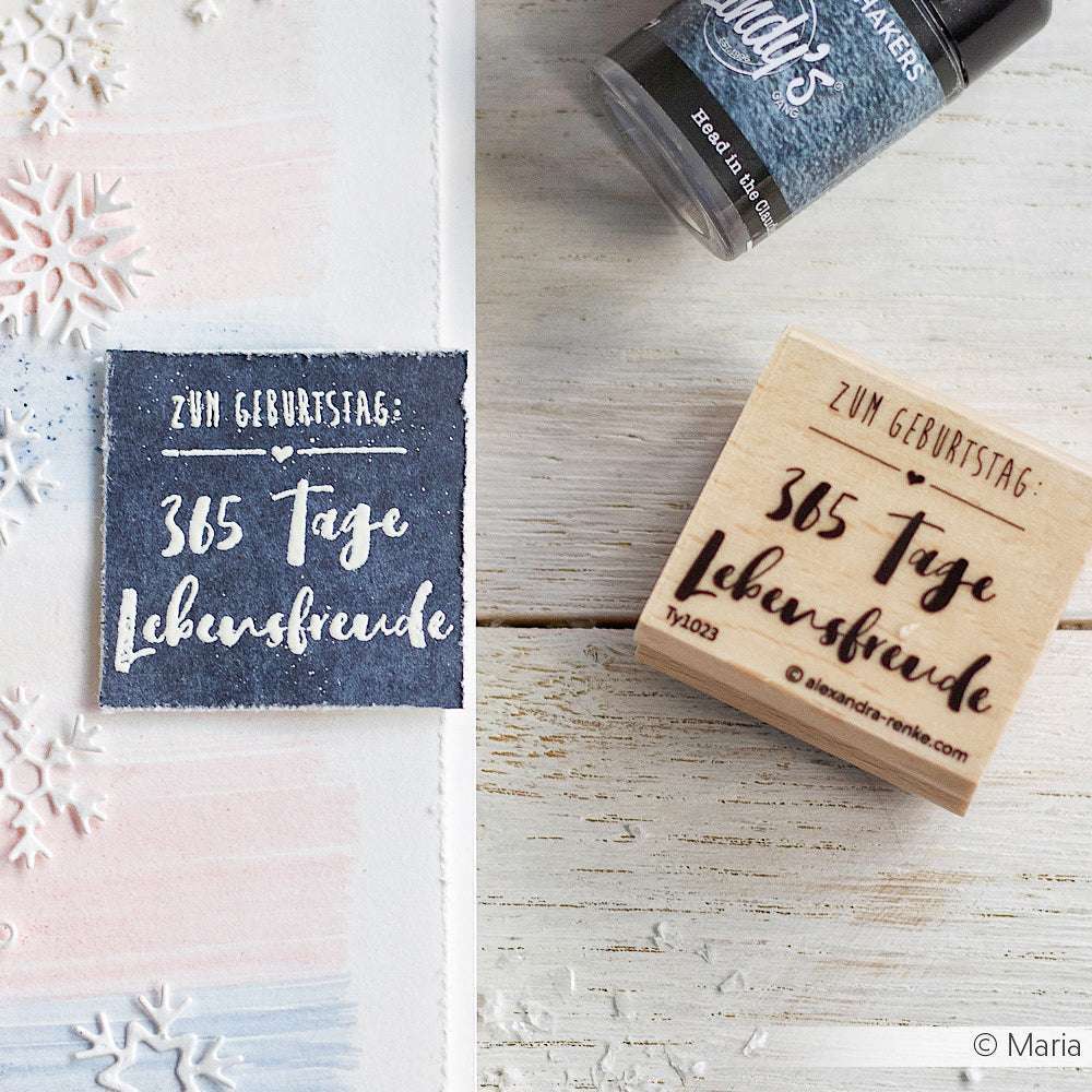 Ein Holzstempel von Alexandra Renke und Tinte mit dem deutschen Text "365 Tage Lebensfreude" auf einer Karte mit Schneeflockenmotiven.