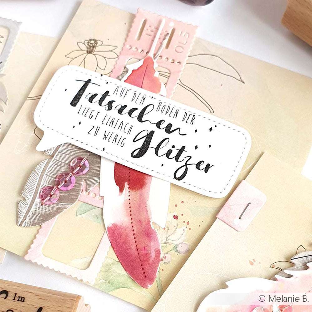 Handgefertigte Karte von Alexandra Renke mit deutscher Kalligrafie, einer rosafarbenen Federillustration, dekorativen Stempelgummi-Stempeln und funkelnden Pailletten.