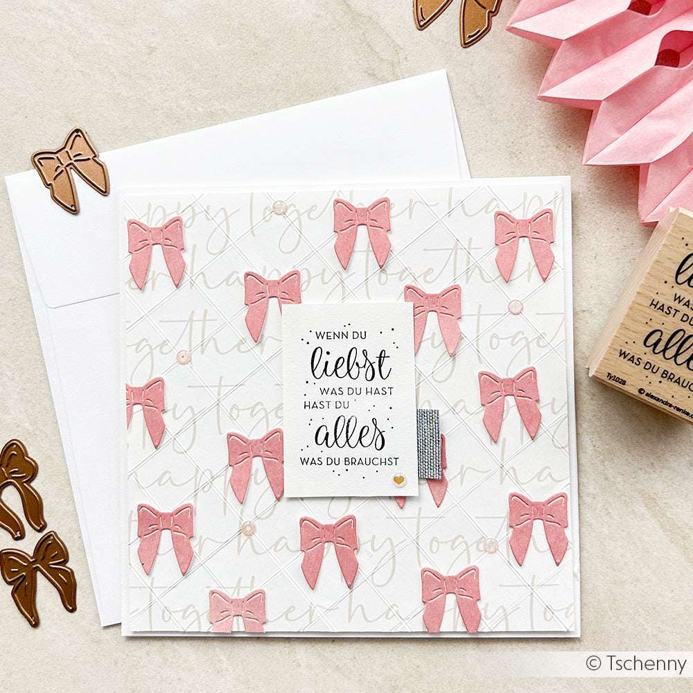 Eine quadratische Alexandra Renke Karte mit rosa Schleifen, einem deutschen Zitat und Stempelmotiv sitzt auf einem Umschlag. In der Nähe befinden sich schleifenförmige Stanzteile und ein charmanter Alexandra Renke Holzstempel für zusätzliche Details.