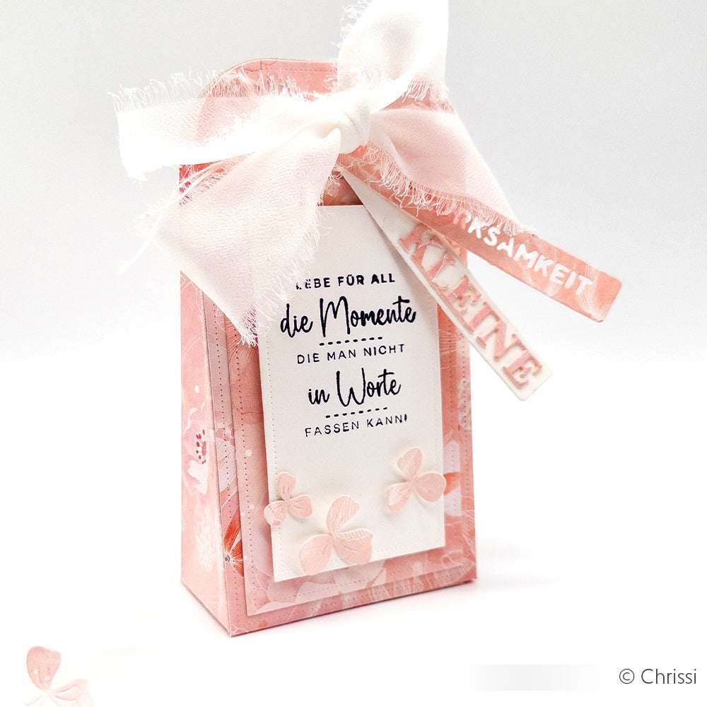 Diese rosafarbene Geschenkschachtel von Alexandra Renke ist mit einer weißen Schleife, floralen Akzenten und einem deutschen Text versehen - perfekt für Papierprojekte oder zur kreativen Gestaltung mit Holzstempeln.