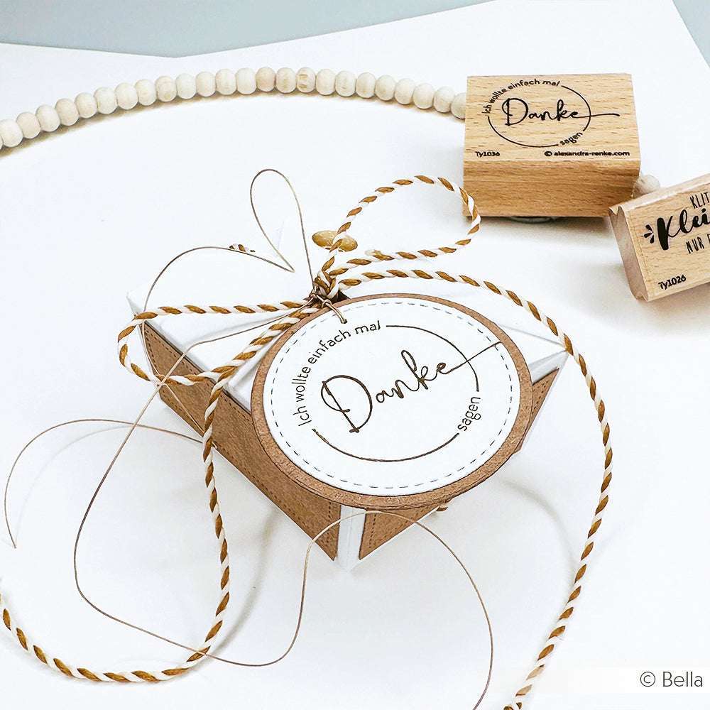 Alexandra Renke-Geschenkbox mit rundem Danke-Anhänger, Bindfaden und passendem Danke-Stempel neben einem Holzstempel auf einer weißen Fläche.