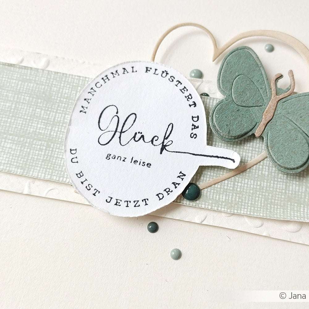 Rundes Etikett mit deutschem Text, einem grünen Schmetterling, blauen Punkten und einem strukturierten blaugrauen Streifen auf Weiß - ideal für Papierprojekte mit Ihrem Lieblingsholzstempel oder Stempelkissen von Alexandra Renke.