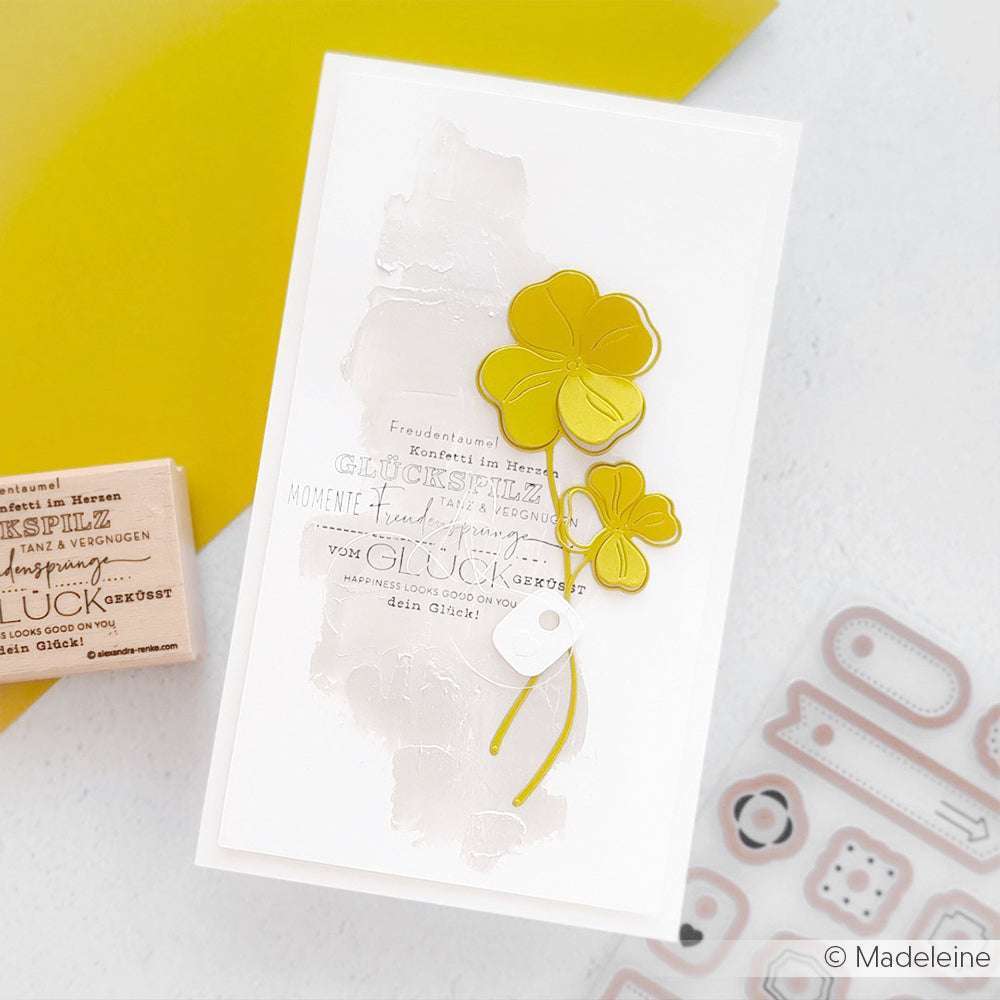 Handgefertigte Karte von Alexandra Renke mit gelben Kleeblumen, deutschem Text und einem weichen beigen Hintergrund mit zarten Glückspilz-Details für ein charmantes Finish. Von oben betrachtet.