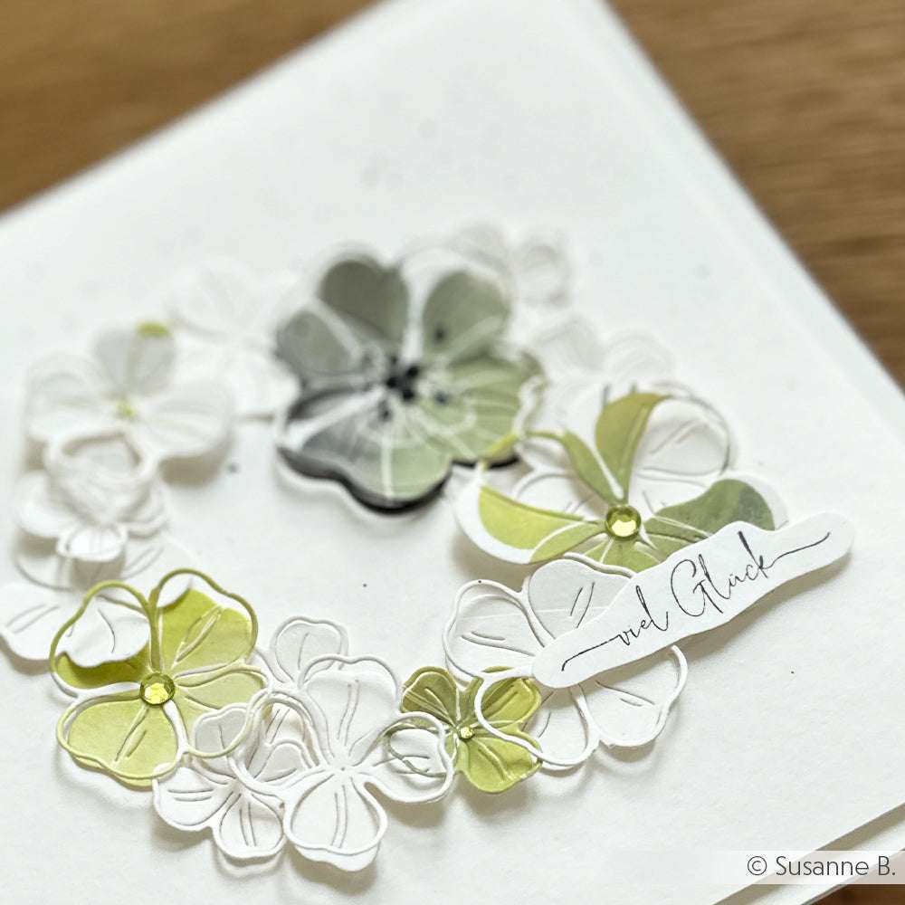 Eine handgefertigte Karte von Alexandra Renke mit zarten grünen, weißen und grauen Papierblumen und einem kleinen, mit dem Spruch "Viel Glück" gestempelten Anhänger.