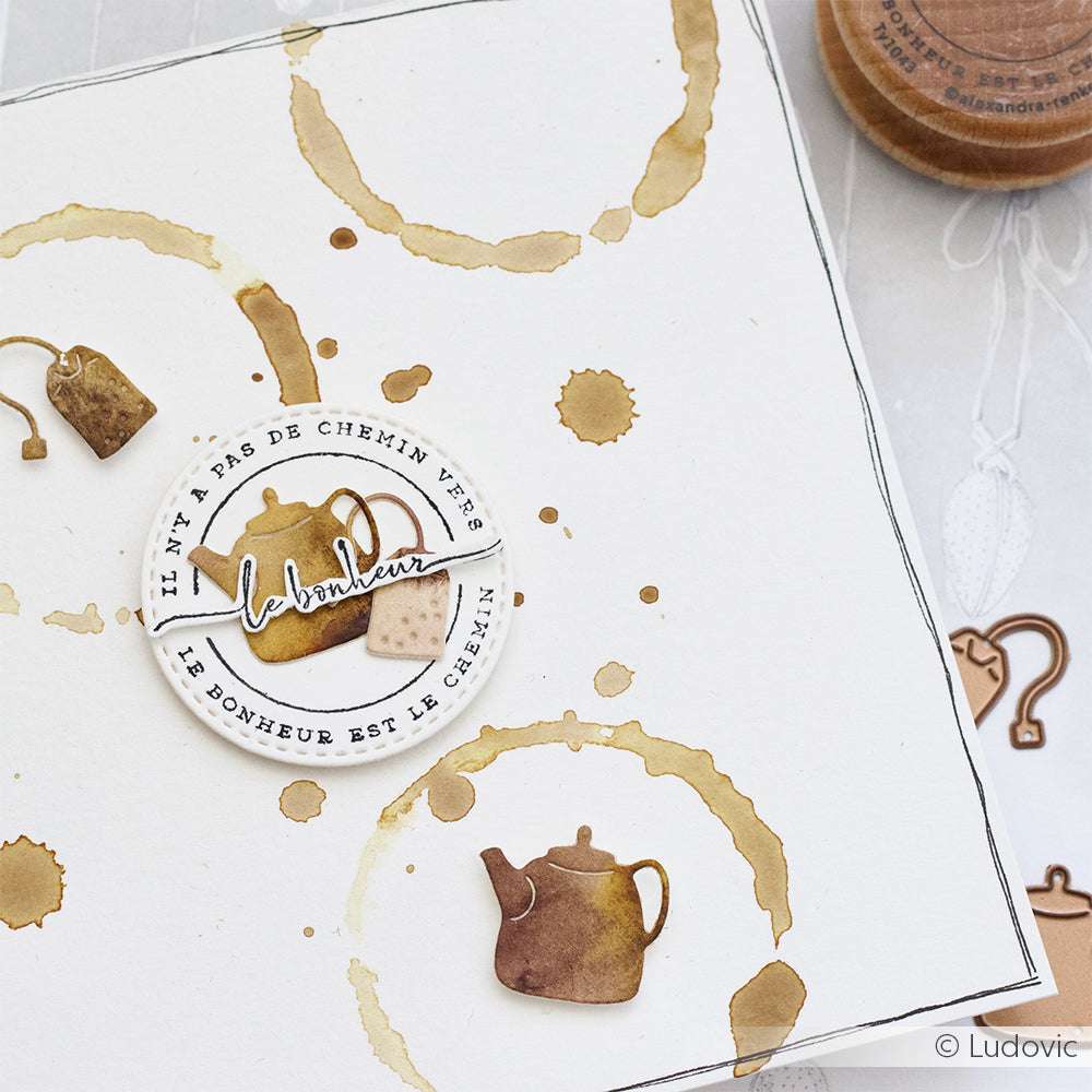 Eine quadratische Karte mit Flecken auf der Kaffeetasse, einer Teekanne, einem Teebeutel und einem französischen Text über das Glück, gestaltet mit Alexandra Renke Motivstempel oder Stempelgummi für charmante Details.