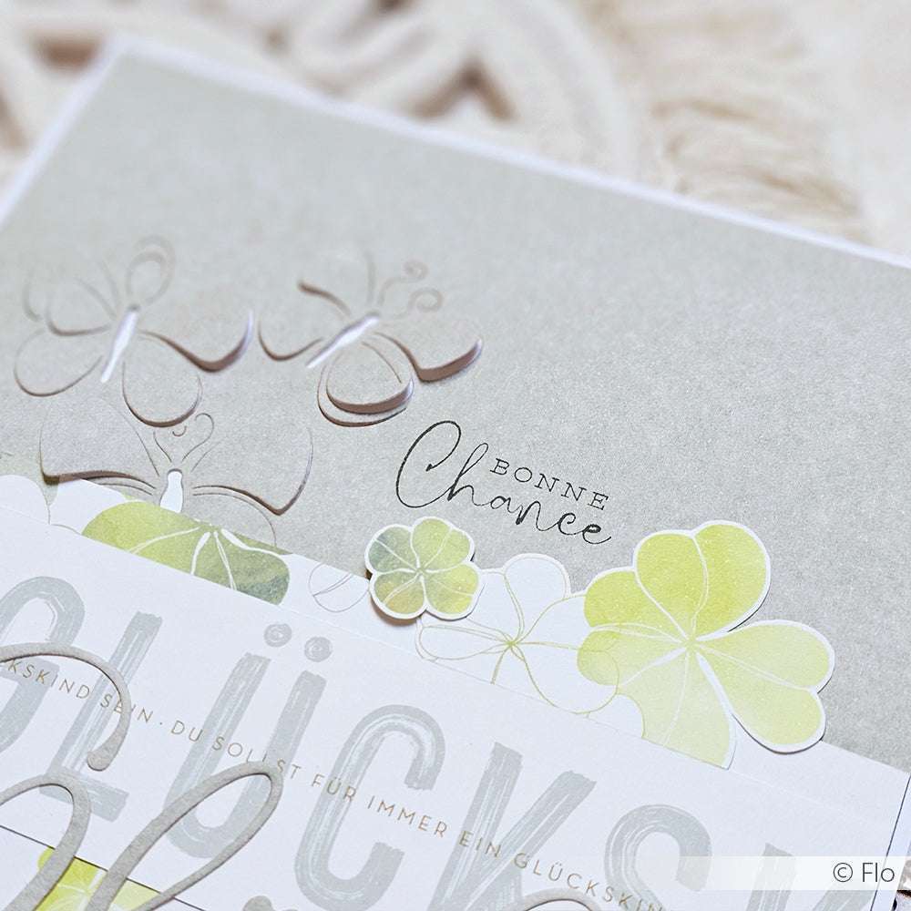 Nahaufnahme einer Karte von Alexandra Renke mit Kleeblättern, Schmetterlingen und dem Text "Bonne Chance" auf einem weichen grauen Hintergrund - ideal für kreative Projekte oder zum Veredeln mit Ihren Lieblingsstempeldesigns.