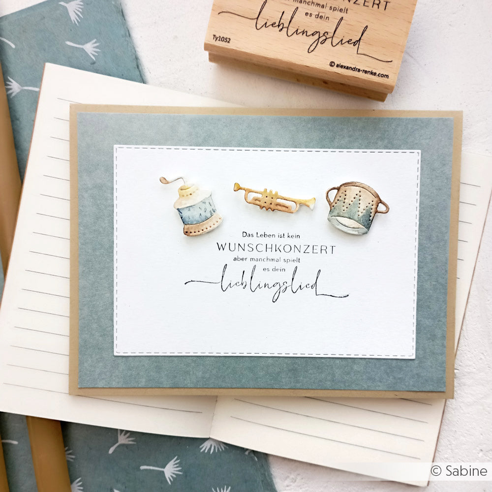 Handgefertigte Alexandra Renke Karte mit einer Spule, Trompete und Sieb über deutschem Text und einem Stempel mit Spruch auf blauem und beigem Hintergrund.