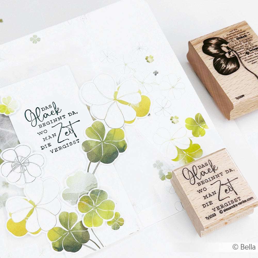 Eine Karte mit grünen Kleemotiven und deutschem Text von Alexandra Renke liegt neben zwei dekorativen Holzstempeln auf einer weißen Fläche, perfekt für Papierprojekte.