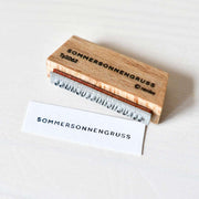 Ein Holzstempel von Alexandra Renke mit der Aufschrift SOMMERSONNENGRUSS und gestempeltem Etikett auf weißem Untergrund - ideal für kreative Papierprojekte.