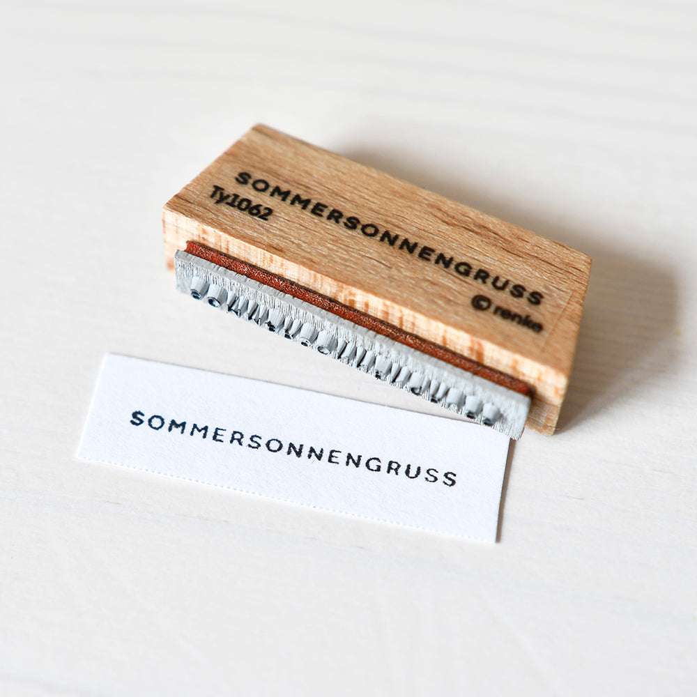 Ein Holzstempel von Alexandra Renke mit der Aufschrift SOMMERSONNENGRUSS und gestempeltem Etikett auf weißem Untergrund - ideal für kreative Papierprojekte.