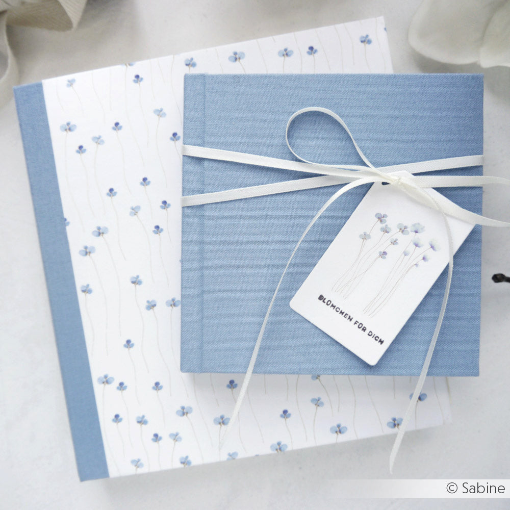 Zwei blau-weiß geblümte Notizbücher von Alexandra Renke, eines mit weißem Band und passendem Anhänger, liegen auf einer hellen Unterlage - perfekt, um sie mit Ihren Lieblingsaccessoires von Holzstempel oder Mini Typo zu kombinieren.