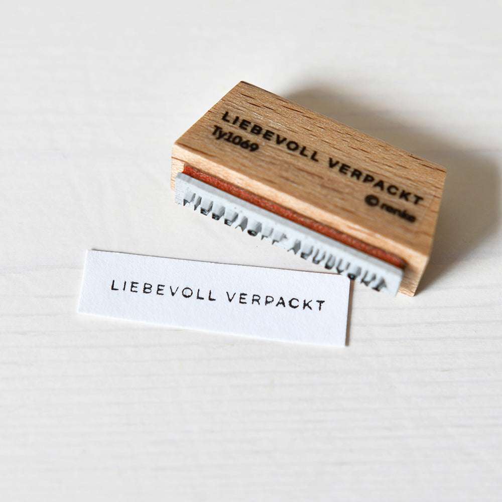 Ein Holzstempel und eine gestempelte Karte von Alexandra Renke mit dem Motiv "LIEBEVOLL VERPACKT" liegen auf weißem Untergrund, daneben befindet sich ein passendes Stempelkissen.