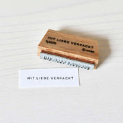 Ein Holzstempel und ein gestempeltes Papier, beide mit dem Motiv "MIT LIEBE VERPACKT" von Alexandra Renke, liegen auf einer hellen Holzfläche neben einem Stempelkissen.