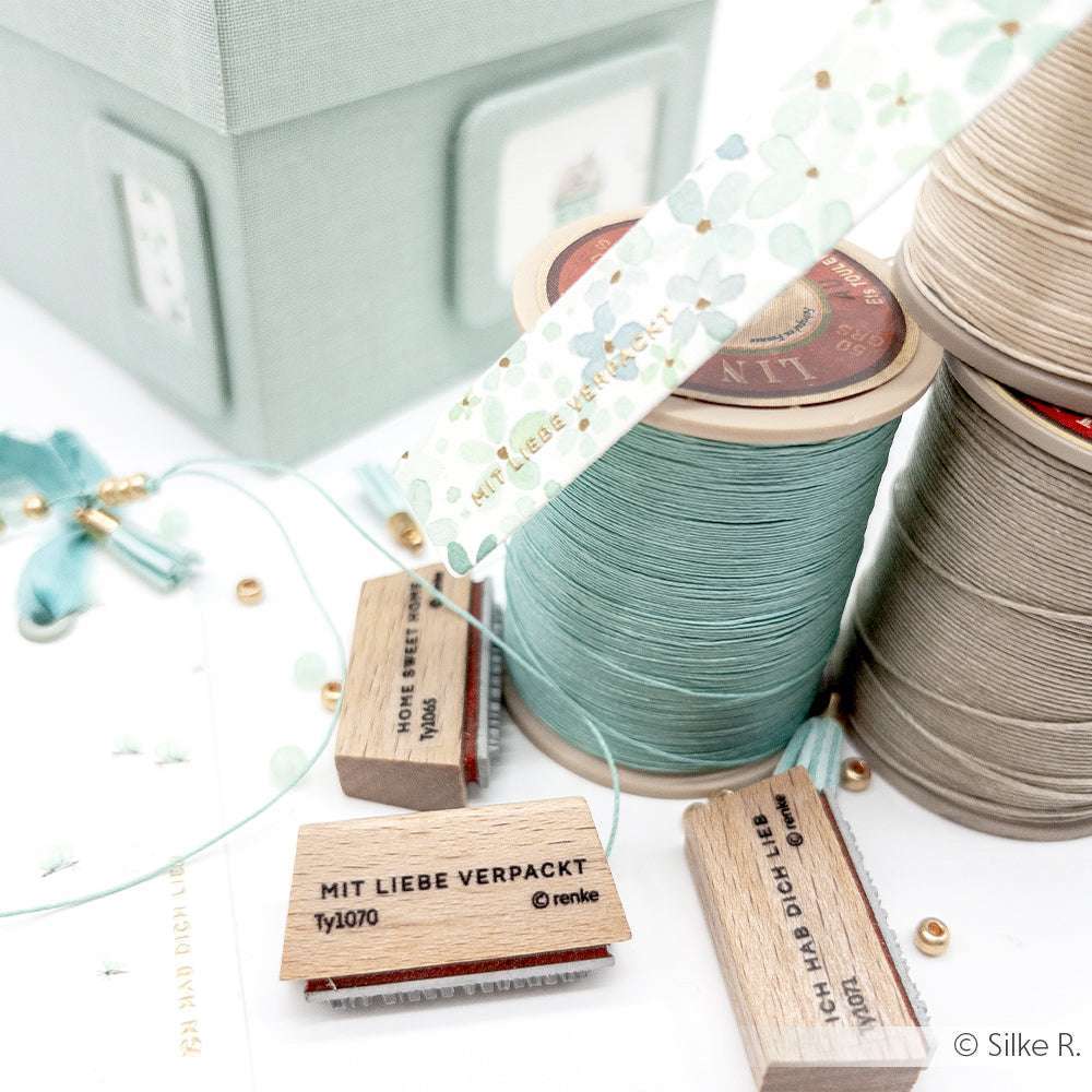 Alexandra Renke hat Spulen mit grünem und beigem Garn, Holzstempel, Perlen und Blumenband auf einer weißen Fläche angeordnet.