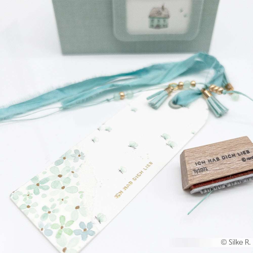 Ein geblümtes Lesezeichen von Alexandra Renke mit tealfarbenen Quasten, ein Holzstempel und eine blaue Karte mit einer Hausillustration.