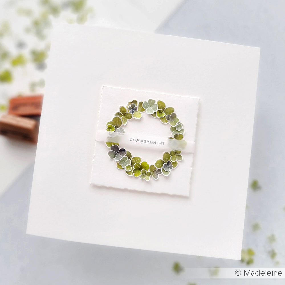 Eine weiße Karte von Alexandra Renke zeigt einen zart gestempelten grünen Blätterkranz mit dem Wort "Glücksmoment" in der Mitte.