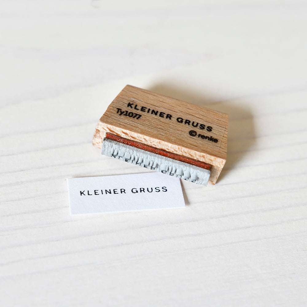 Ein Holzstempel von Alexandra Renke mit der Aufschrift KLEINER GRUSS liegt neben einem gestempelten Text auf weißem Papier, der den Stil der Mini Typo unterstreicht.