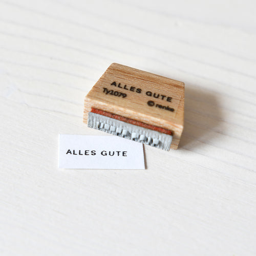 Ein Alexandra Renke Holzstempel mit "ALLES GUTE" und ein gestempeltes Papier mit demselben Spruch liegen auf einer weißen Fläche - ideal für Mini-Typo-Paper-Projekte.