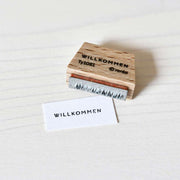 Ein Alexandra Renke Holzstempel und Papier zeigen beide "WILLKOMMEN" auf einer hellen Oberfläche - ideal für Papierprojekte oder als Mini-Typo-Akzent für Ihre Bastelarbeiten.