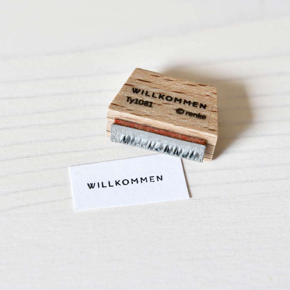 Ein Alexandra Renke Holzstempel und Papier zeigen beide "WILLKOMMEN" auf einer hellen Oberfläche - ideal für Papierprojekte oder als Mini-Typo-Akzent für Ihre Bastelarbeiten.