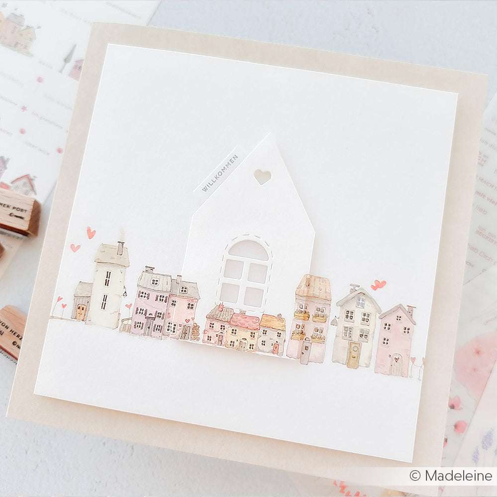 Eine handgefertigte Karte von Alexandra Renke mit pastellfarbenen Häusern in Aquarellfarben, einem weißen Hausausschnitt, kleinen rosa Herzen und dem mit einem Stempelkissen gestempelten Satz "Mini Typo Willkommen".