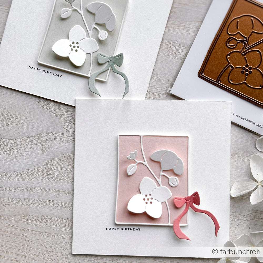 Zwei florale Geburtstagskarten von Alexandra Renke mit 3D-Ausschnittblumen, Schleifen und Pastelltönen ruhen auf einer hellen Holzoberfläche - mit einem "Happy Birthday Stempel" für einen charmanten handgefertigten Look.