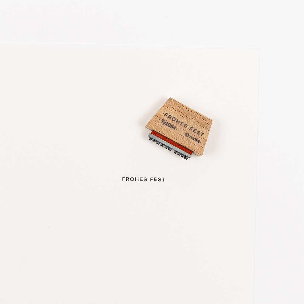 Ein Holzstempel mit Stempelgummi hat den Text „FROHES FEST“ auf ein weißes Blatt Papier gestempelt.