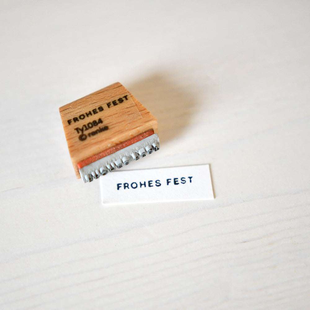 Ein Alexandra Renke Holzstempel mit der Aufschrift Mini Typo Frohes Fest liegt neben weißem Papier, das mit einem festlichen Gruß bestempelt ist - perfekt für die Kombination mit Ihren Lieblingsstempelkissen.