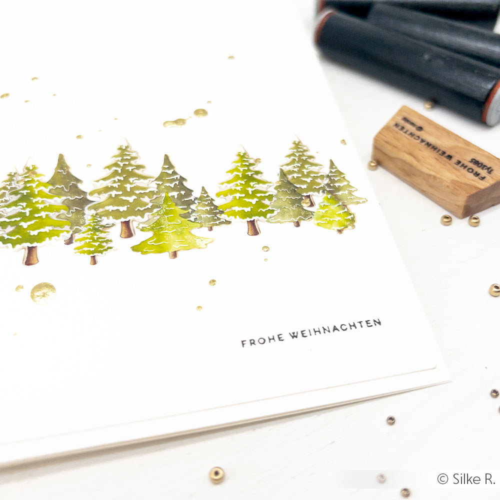 Minimalistische Weihnachtskarte von Alexandra Renke mit grünen Bäumen, goldenen Akzenten und dem Stempel "Frohe Weihnachten" auf weißem Papier - ideal, um Ihren Papierprojekten eine besondere Note zu verleihen.
