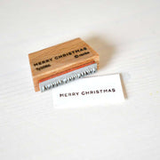 Ein Alexandra Renke Holzstempel und eine gestempelte Karte, beide mit "MERRY CHRISTMAS", sind auf einer weißen Fläche ausgestellt - perfekt, um mit Ihrem Merry Christmas Stempel eine festliche Note zu verleihen.