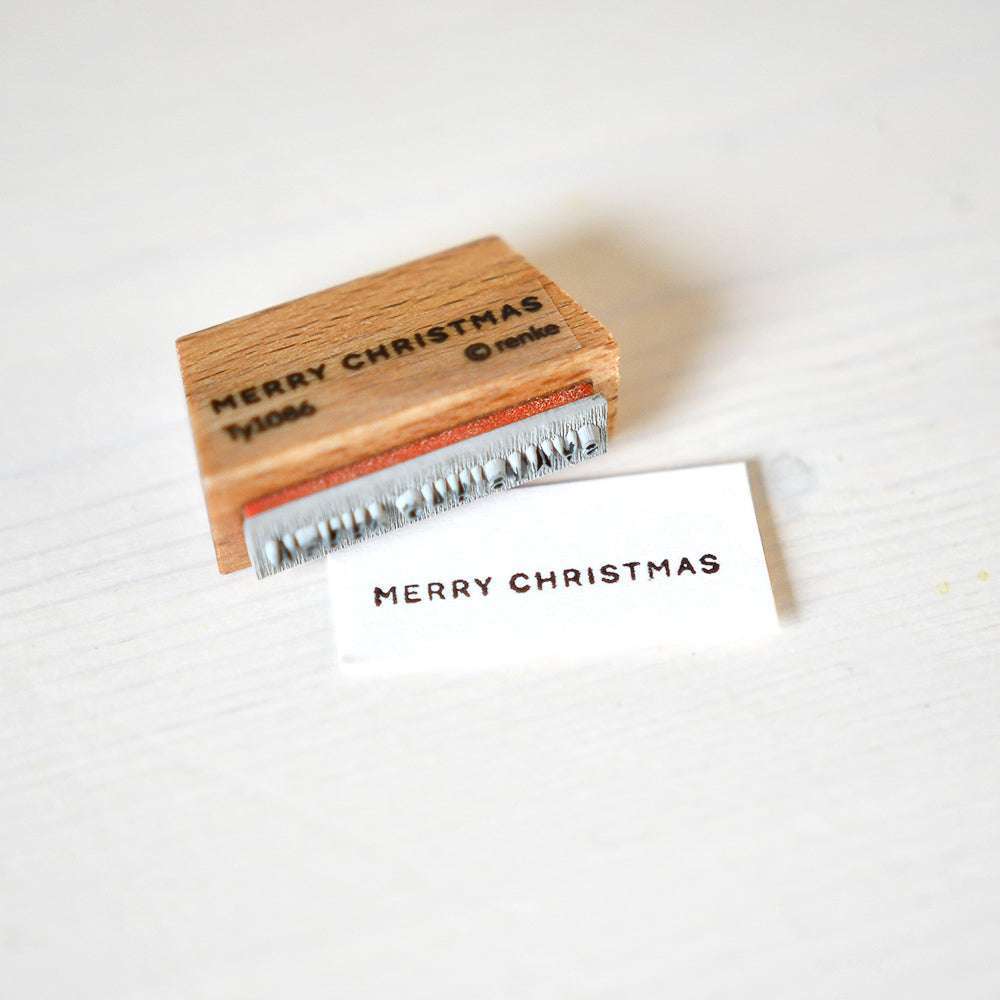 Ein Alexandra Renke Holzstempel und eine gestempelte Karte, beide mit "MERRY CHRISTMAS", sind auf einer weißen Fläche ausgestellt - perfekt, um mit Ihrem Merry Christmas Stempel eine festliche Note zu verleihen.