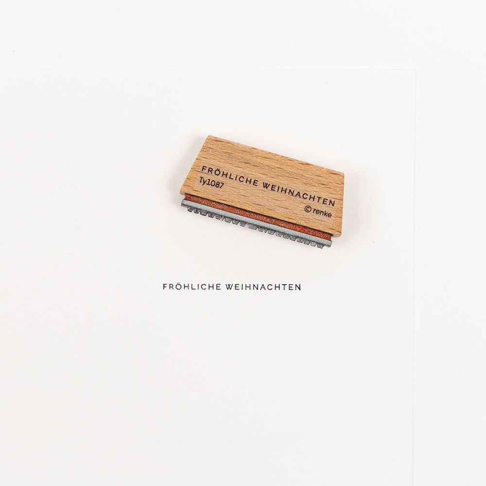 Holzstempel mit Stempelgummi und der Aufschrift „FRÖHLICHE WEIHNACHTEN“ über dem gestempelten Text auf einem leeren weißen Blatt Papier.