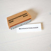 Ein hölzerner Alexandra Renke Holzstempel mit "WEIHNACHTSWUNDER" und ein gestempeltes Stück Papier mit einem Stempelkissen.