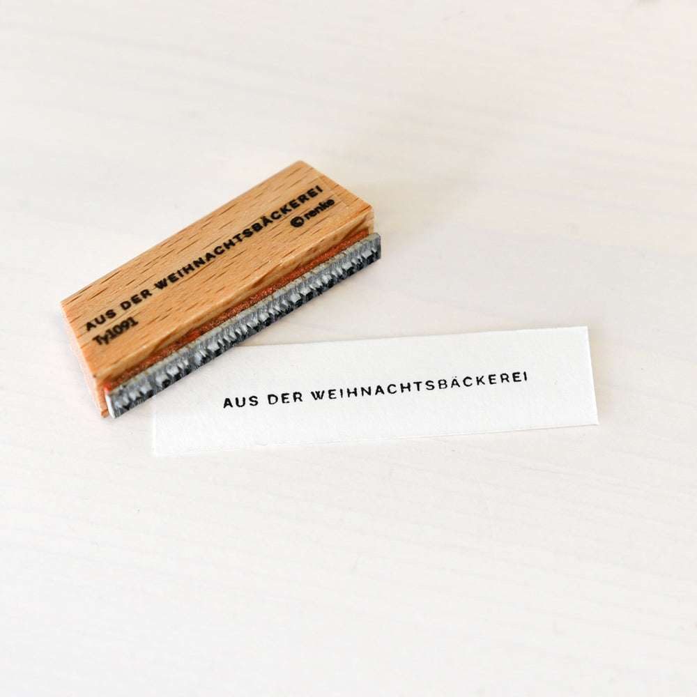 Ein Alexandra Renke Holzstempel und ein mit AUS DER WEIHNACHTSBÄCKEREI gestempeltes Papier ruhen auf einer weißen Oberfläche und verleihen Ihrer Weihnachtsbäckerei festlichen Charme.