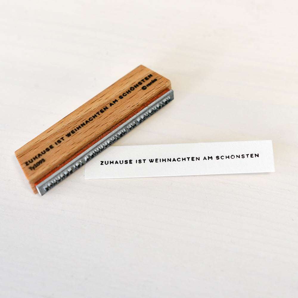 Ein Holzstempel und ein Stempelkissen von Alexandra Renke auf weißem Untergrund, das Papier zeigt den Abdruck: ZUHAUSE IST WEIHNACHTEN AM SCHÖNSTEN.