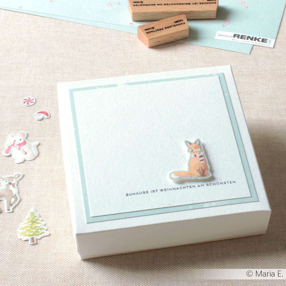 Quadratische Schachtel mit einer Fuchs-Illustration und deutschem Text, umgeben von festlichen Aufklebern, Stempelkissen und Holzstempel-Designs von Alexandra Renke - ideal, um Ihre Weihnachtsstempel-Sammlung zu erweitern.