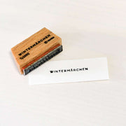 Ein Holzstempel von Alexandra Renke mit Stempelgummi und Papierstempel WINTERMÄRCHEN ruht auf einer hellen Fläche und versprüht den Charme handwerklicher Eindrücke.