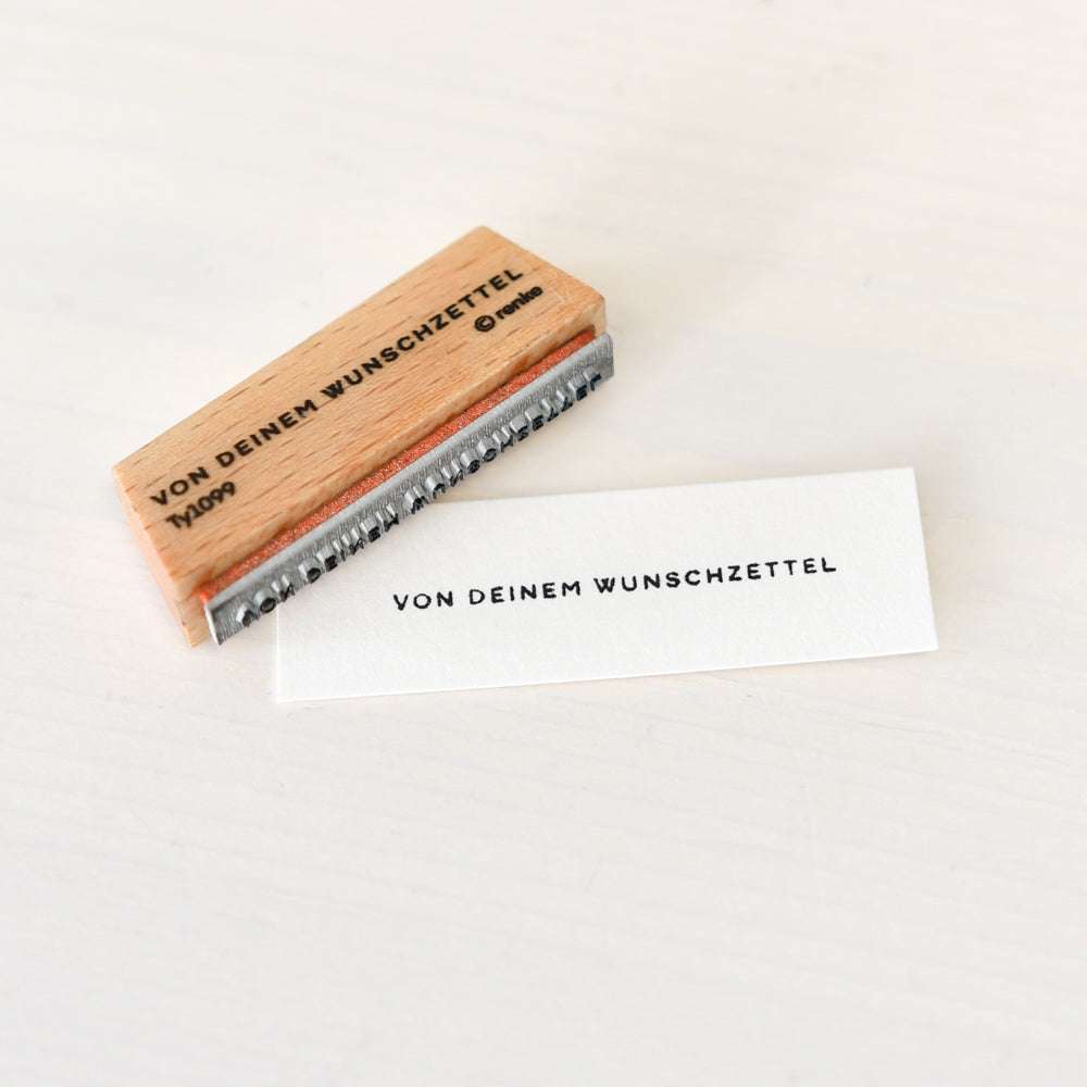 Ein Holzstempel und gestempeltes Papier von Alexandra Renke mit "VON DEINEM WUNSCHZETTEL" liegen auf weißem Grund - ideal für kreative Papierprojekte.
