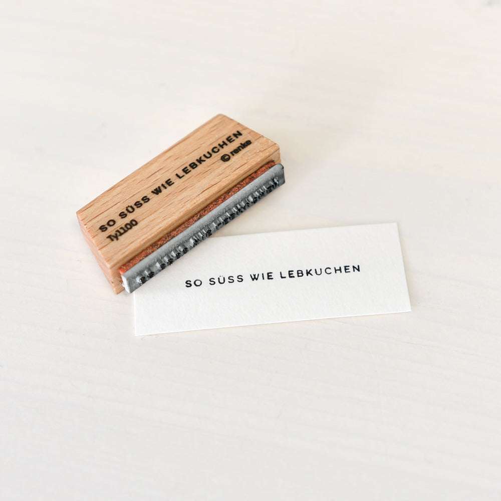 Ein Mini Typo Holzstempel von Alexandra Renke und mit "SO SÜSS WIE LEBKUCHEN" bedrucktes Papier sind auf einer weißen Fläche ausgestellt.