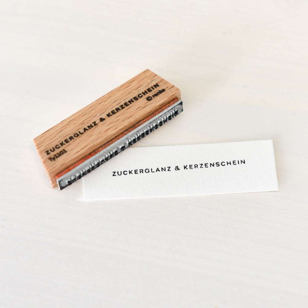 Ein Holzstempel und gestempeltes Papier von Alexandra Renke mit der Aufschrift ZUCKERGLANZ & KERZENSCHEIN auf weißer Fläche - ideal für kreative Papierprojekte.
