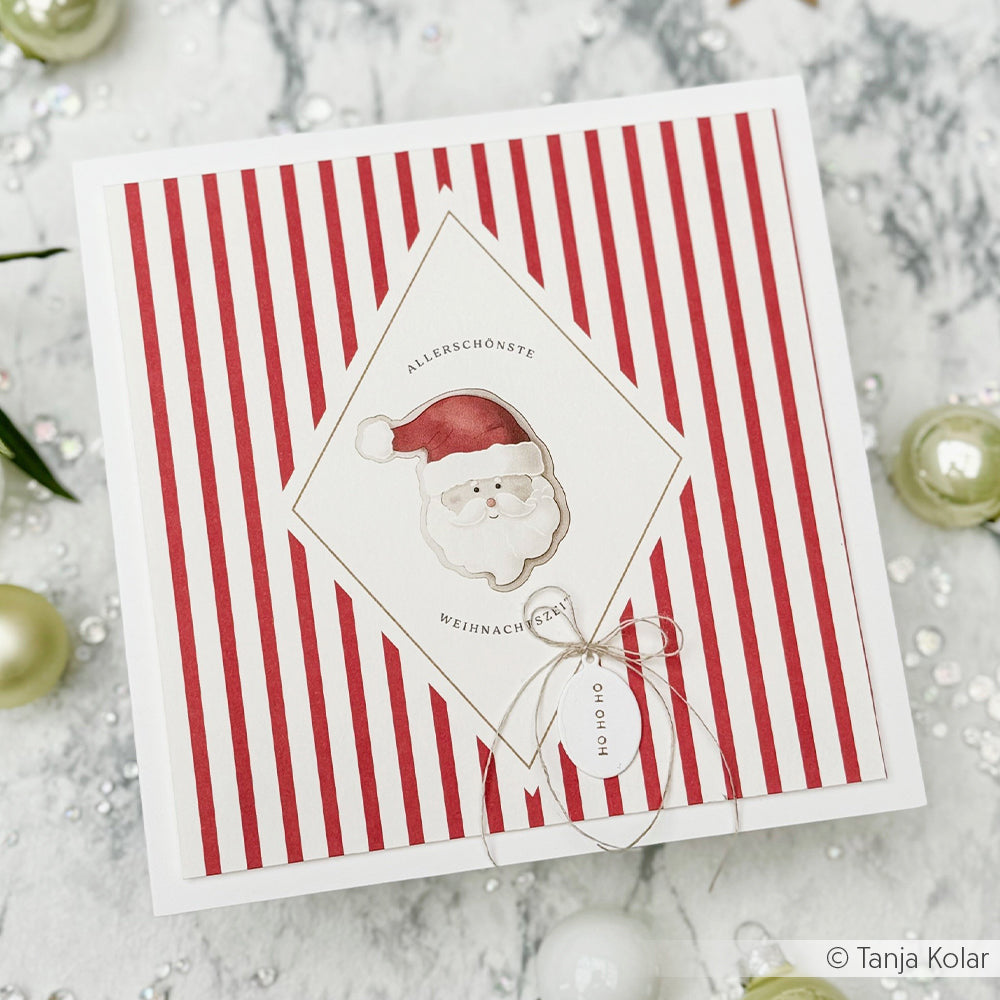 Handgefertigte Weihnachtskarte mit einer Illustration eines Weihnachtsmanngesichts, einem rot gestreiften Hintergrund und dem Text Allerschönste Weihnachtszeit, der mit einem Holzstempel aufgebracht wurde.