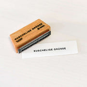 Ein Holzstempel von Alexandra Renke mit dem Motiv "KUSCHELIGE GRÜSSE" und Abdruck auf weißem Papier, ideal in Kombination mit einem passenden Stempelkissen.