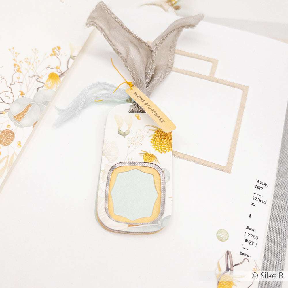 Eleganter handgefertigter Geschenkanhänger mit geschichteten Rahmen und einer Schleife, erstellt von Alexandra Renke, auf einem floralen Kartenhintergrund.