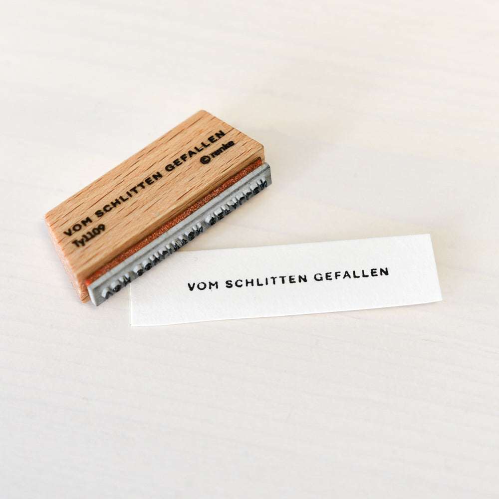 Ein hölzerner Stempel von Alexandra Renke und gestempeltes Papier mit der Aufschrift VOM SCHLITTEN GEFALLEN auf einer hellen Oberfläche, die einen scharfen Mini Typo-Aufdruck zeigt.